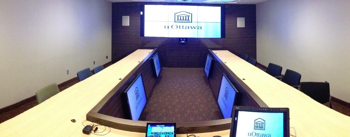 A modern conference room with a large U-shaped table, surrounded by chairs and equipped with multiple laptops, and a projector screen displaying the “uOttawa” logo