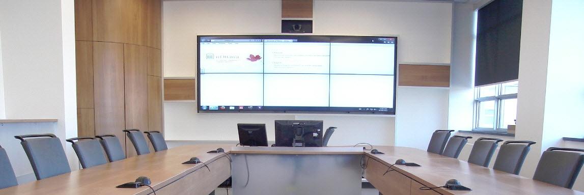 A modern conference room with a long table, multiple chairs, and a large dual-screen presentation setup on the wall