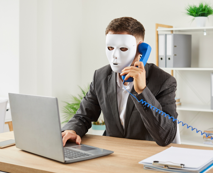 Masked man wearing a suit on the phone while using a computer on a desk behind him a cabinet file r