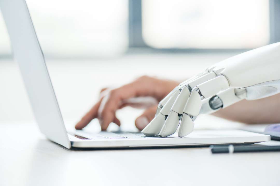 robotic hand next to a human hand typing in a white laptop 