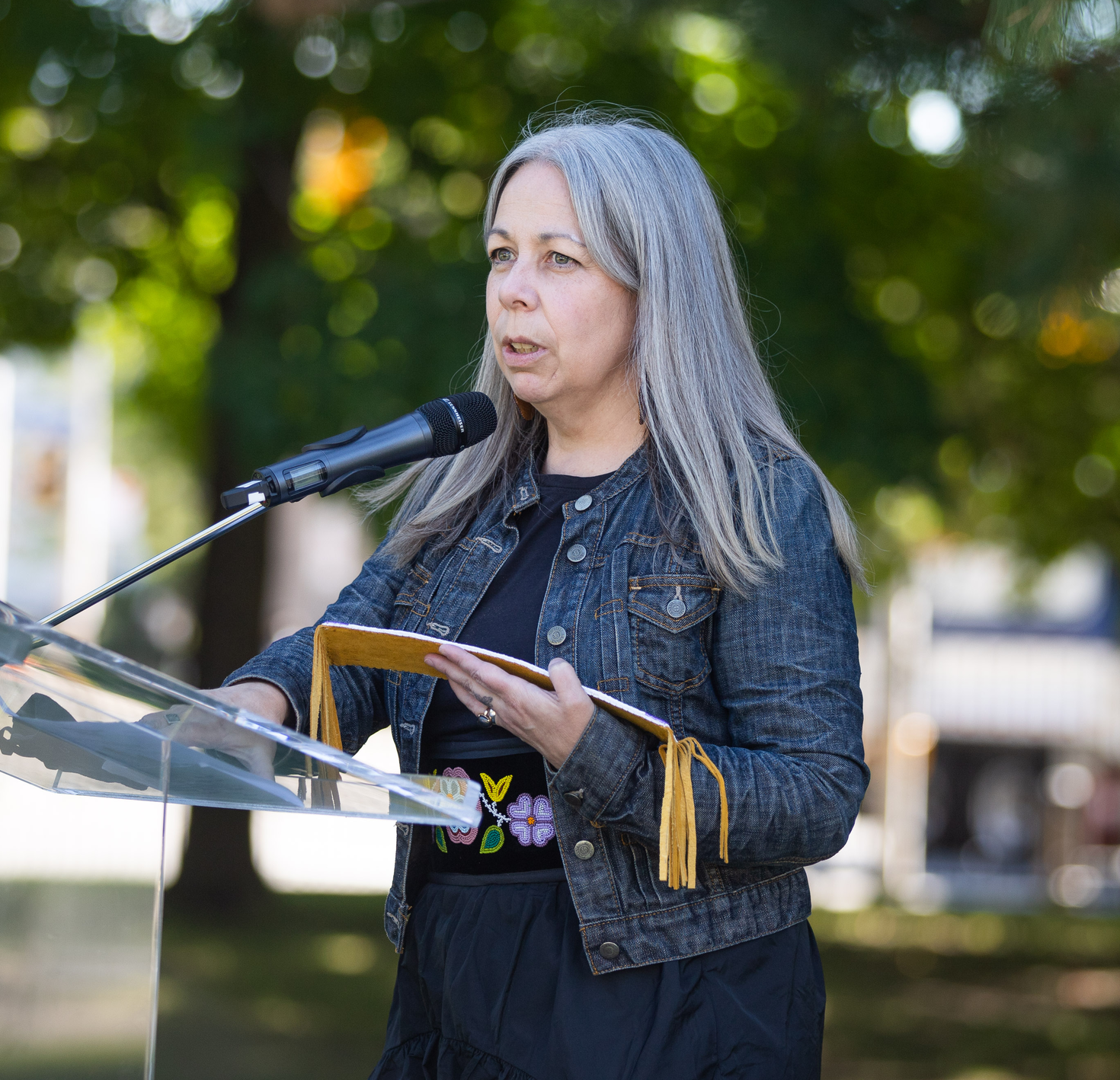 Image shows Dr. Brenda Macdougall, a Métis woman, speaking outside at a podium with a blurred background of trees behind her.