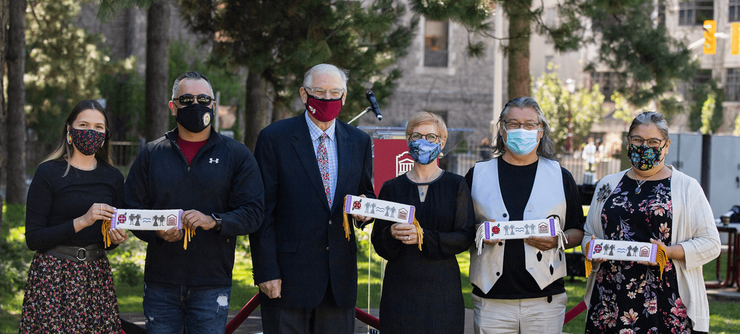 Six masked individuals standing side by side. Four of them are holding beaded belts meant to signify a mutual understanding and respect.