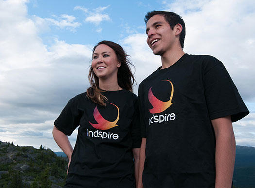 Two students, both wearing black Indspire t-shirts, looking to the left. A blue sky and some trees are visible in the background.