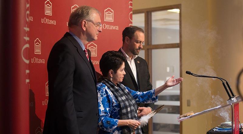 Profile shot of three individuals, a woman in the center and two men to her sides, standing together behind a podium. The woman is holding a feather while she points to the right of the image.