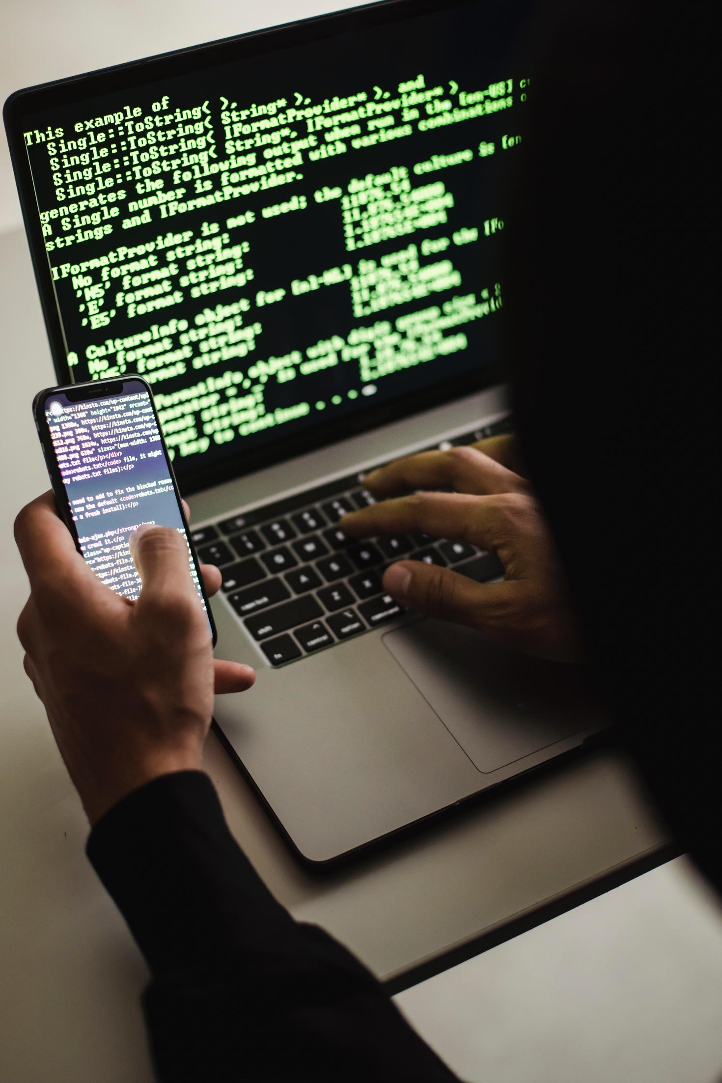 Individual at laptop performing a cyber attack with cellphone in hand