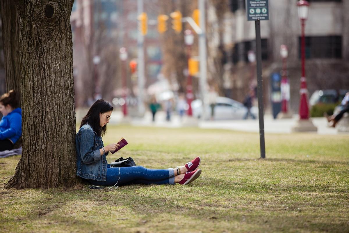Student sitting against tree reading a book on Tabaret lawn