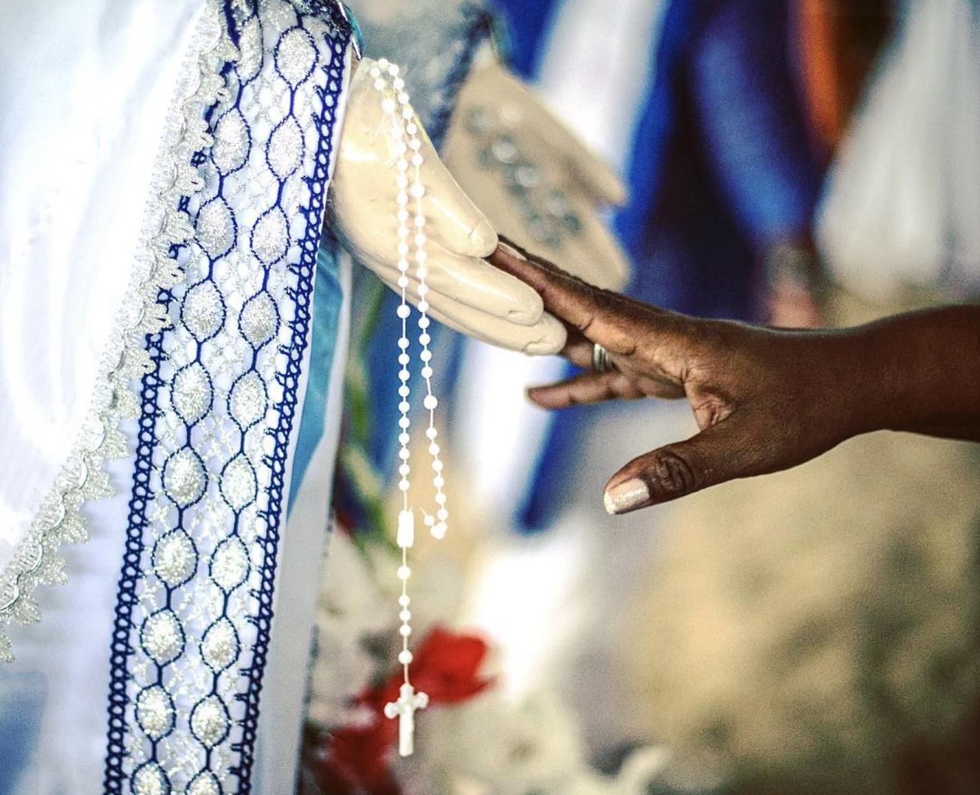 Person reaching out to touch the hand of a religious statue holding beads