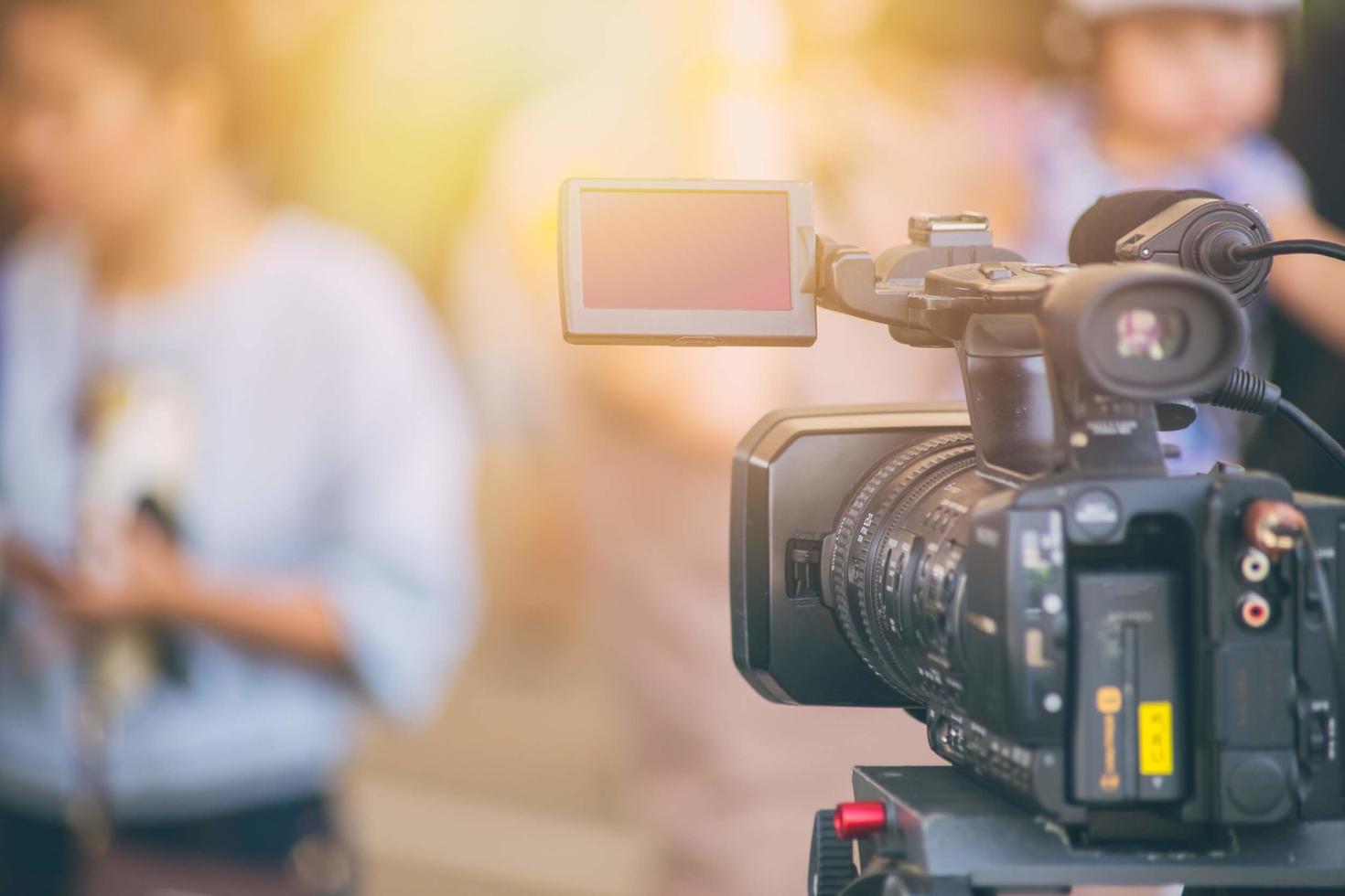 A professional video camera on a tripod focused in the foreground with a blurred background featuring an individual holding a microphone