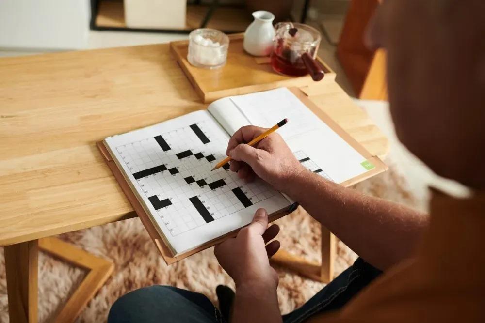 an individual seated at a table, holding a pen and completing a crossword game on paper