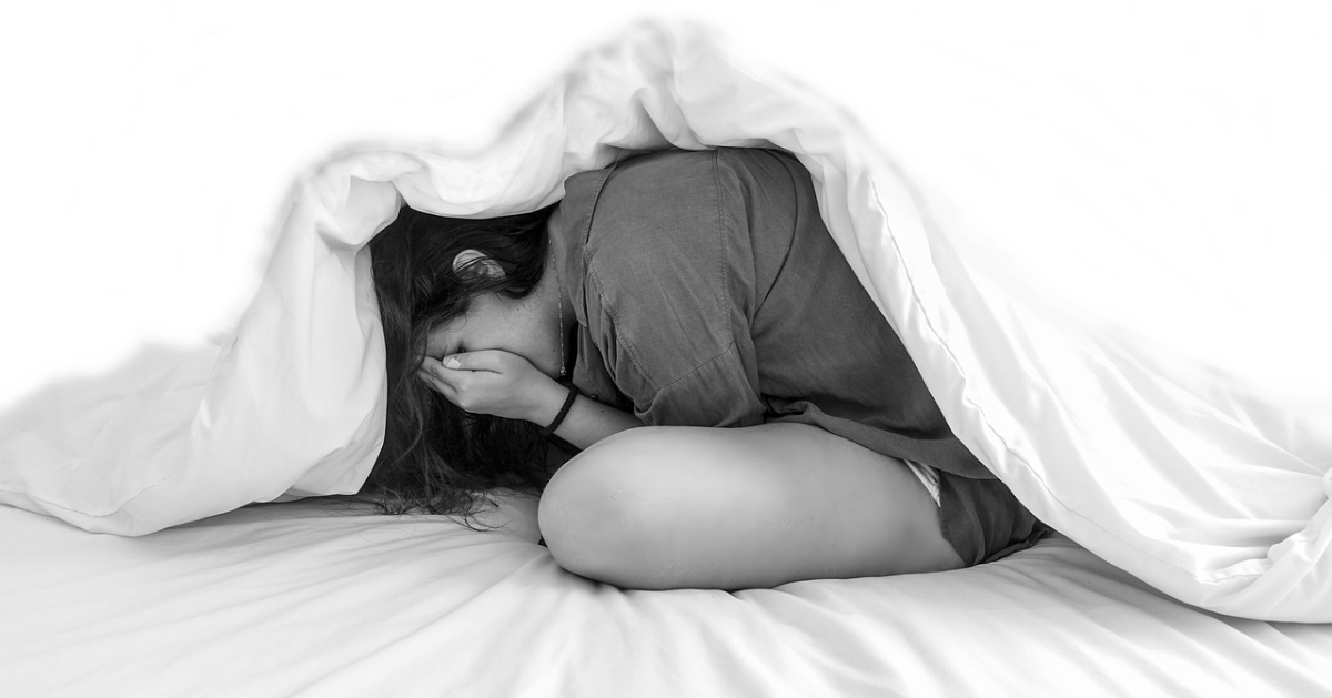 A young woman under her bed covers hides her face in her hands