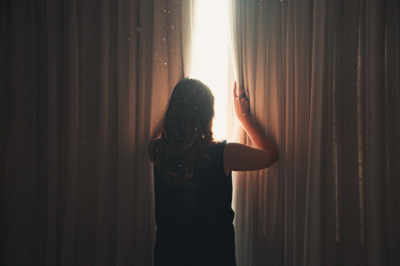 Woman in dark room peaking through blinds to outside