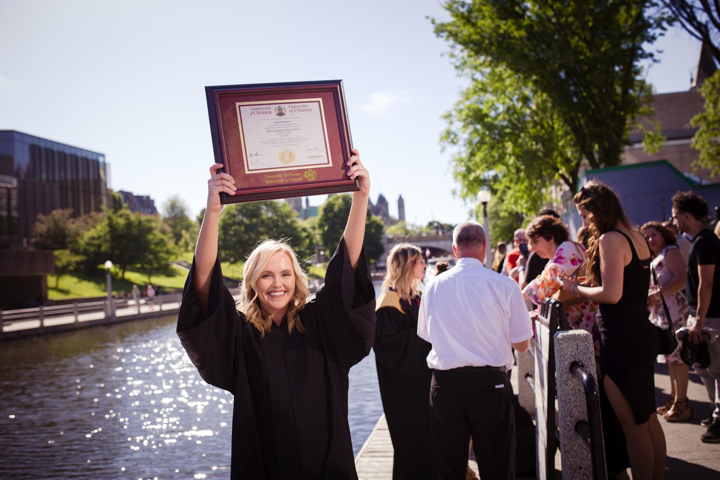 uOttawa graduate holding up framed diploma beside the canal.