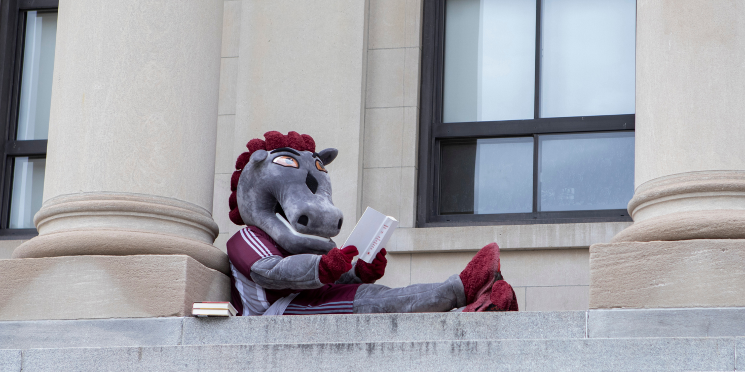 Gee-gees mascot reading book outside of building on campus.