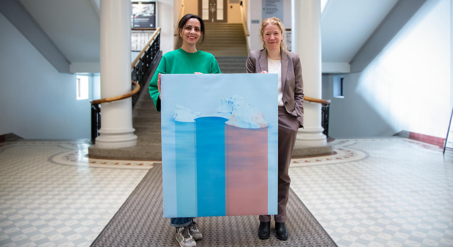 Mina Moshajjari, student in the Master of Fine Arts program, and Julie St-Pierre, interim vice-president, Research and Innovation, holding Mina’s winning piece “Nature’s response”, in the lobby of the University of Ottawa's Tabaret building, holding Mina's winning work, ‘Nature's response’. The work depicts an iceberg with a view above and below the water, with pink and blue hues.