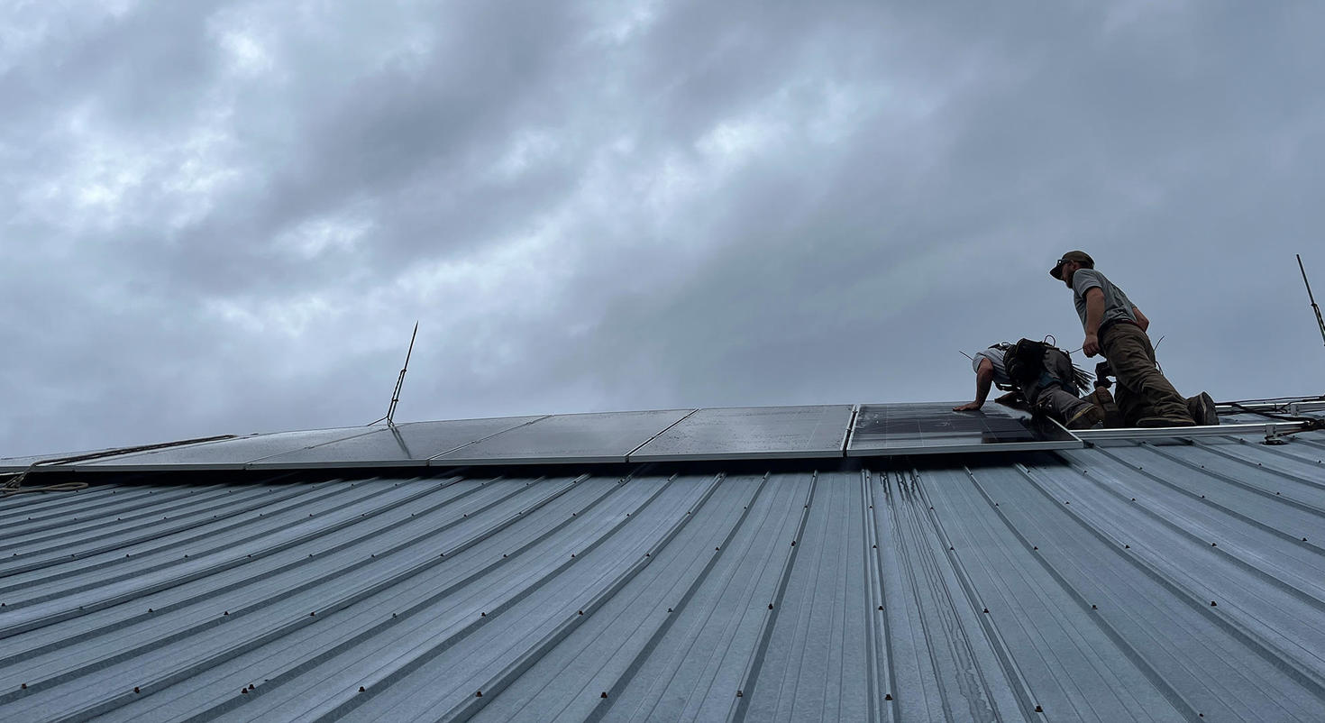 Two workers installing solar panels on a metal roof under a cloudy sky