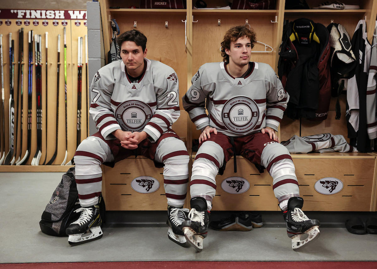 Two Gee-Gees men's hockey players sitting in the locker room, wearing Telfer night jerseys