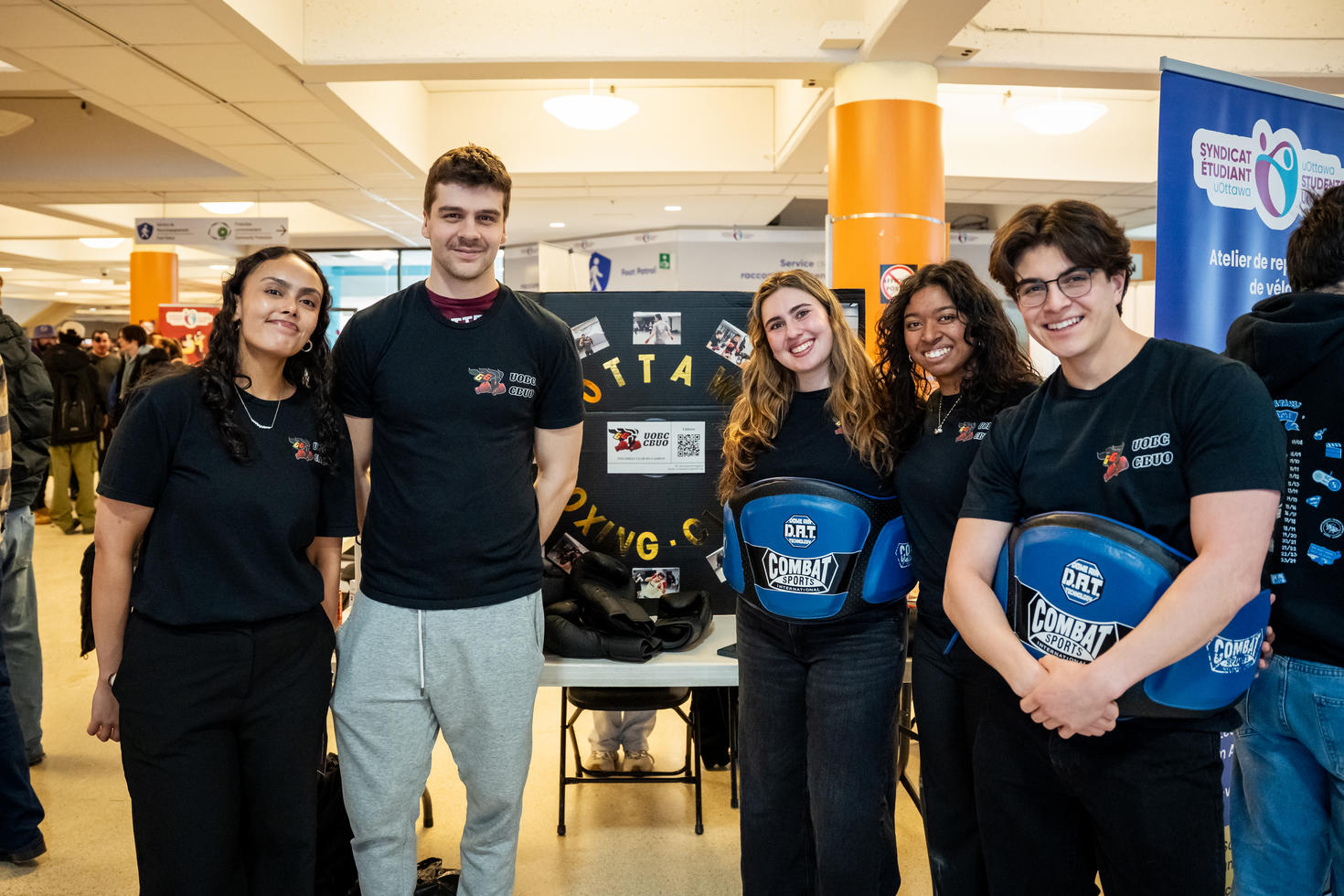 uOttawa Boxing Club executives standing in front of their club display at the Winter Clubs Fair. Two wear blue protective gear around their waists. A black tri-fold board behind them features the club name in gold letters, a logo, a QR code, and photos of boxing.