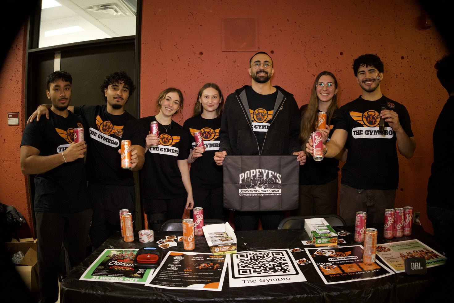 The GymBro executives at the Winter Clubs Fair, standing behind a table covered in GURU energy drinks, a QR code, stickers, bars, and flyers. The seven members wear matching black t-shirts with the club logo and hold up cans of energy drinks, while the member in the center holds a Popeye's Supplements banner.