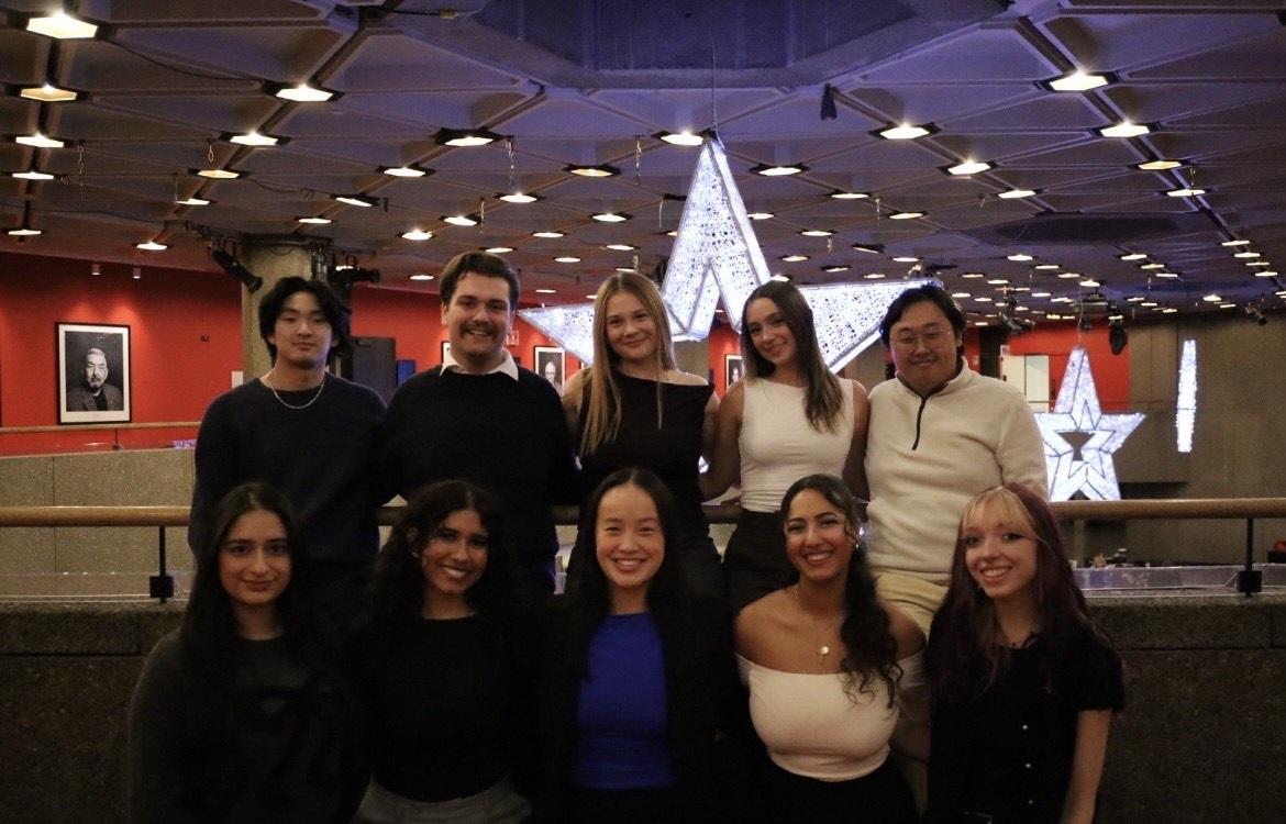 Group photo of the uOttawa Multiple Sclerosis Club executive team, standing and kneeling indoors, smiling at the camera. They are in a modern venue with red walls, framed portraits, and large illuminated star decorations in the background.