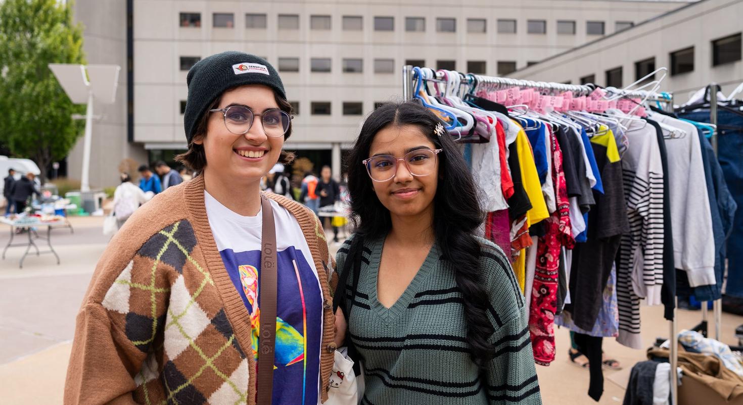 Two people smiling at the camera at an outdoor Free Store Pop-Up, standing in front of clothing racks filled with donated clothes.