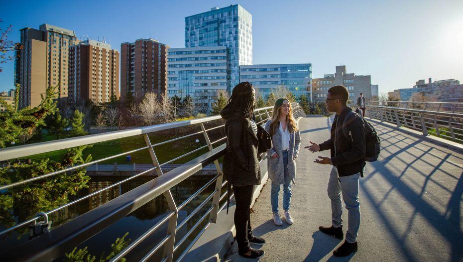 three students talking on Ottawa pedestrian bridge in front of Faculty of Social Sciences building