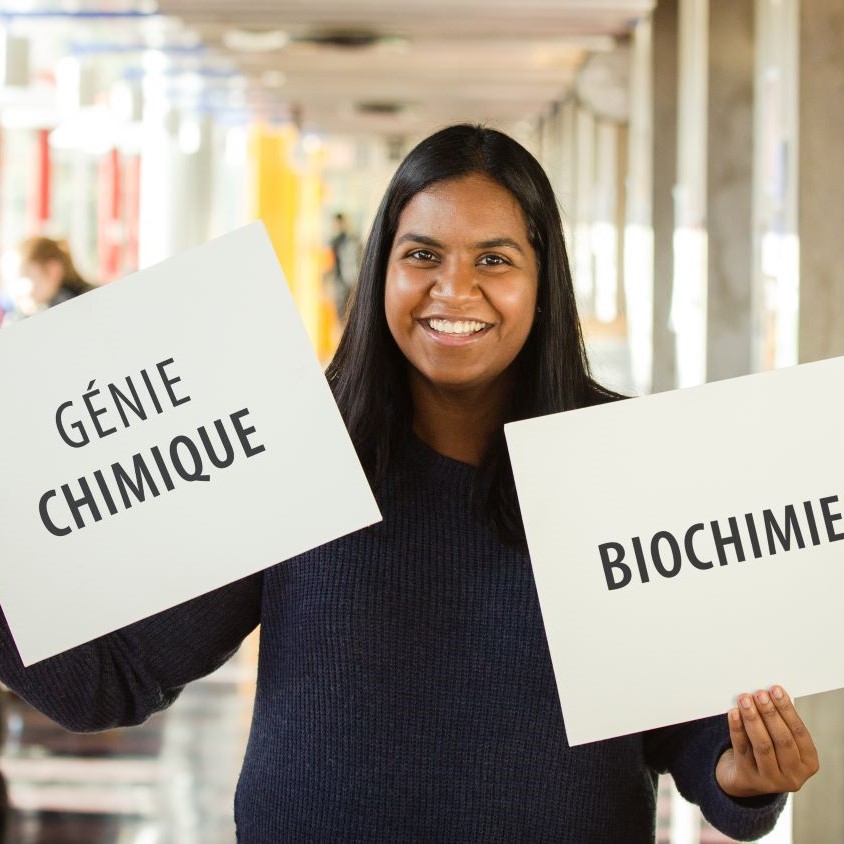 une étudiante qui tient deux affiches sur lesquelles sont écrit Génie chimique et Biochimie