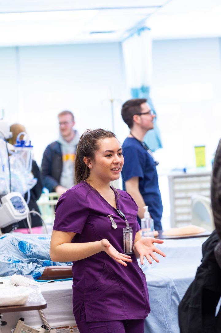 Une étudiante infirmière en blouse d'hôpital accueille les participants à une journée portes ouvertes, un étudiant infirmier se tient à l'arrière-plan.