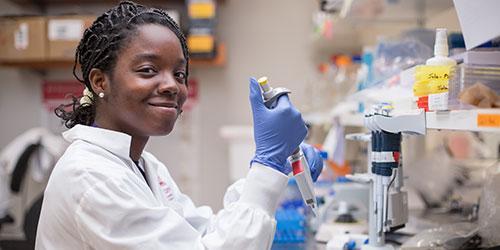 Une étudiante vêtue d’une blouse de laboratoire sourit en travaillant avec des éprouvettes dans un laboratoire.
