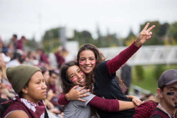 Two students with garnet and white stripes painted on their faces hug and cheer at a sporting event.