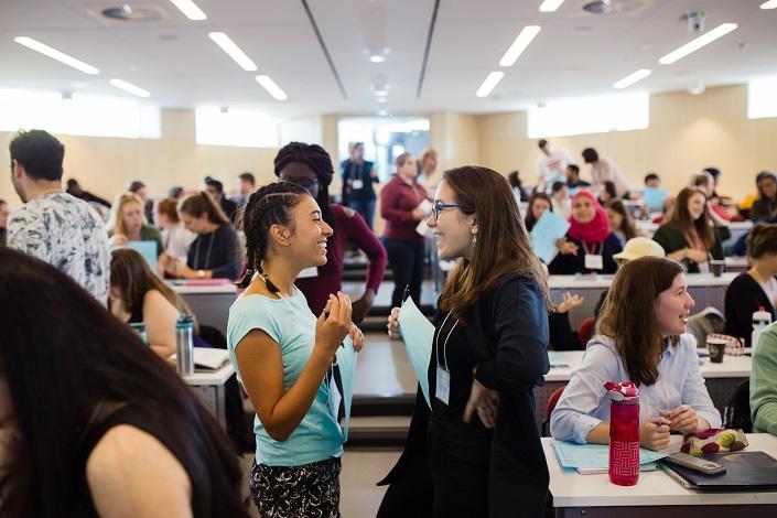 Deux étudiantes souriantes échangent en face à face devant la classe; d’autres étudiants en arrière-plan échangent aussi en groupes.