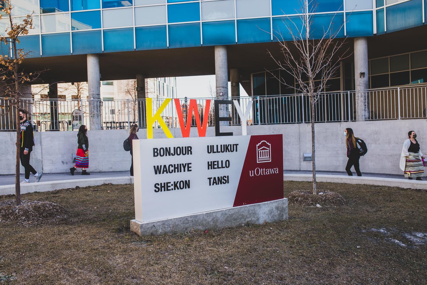 Étudiants autochtones qui marchent le long d'un chemin derrière le panneau qui dit KWEY sur le campus, le panneau aussi dit bonjour dans des différentes langues autochtones