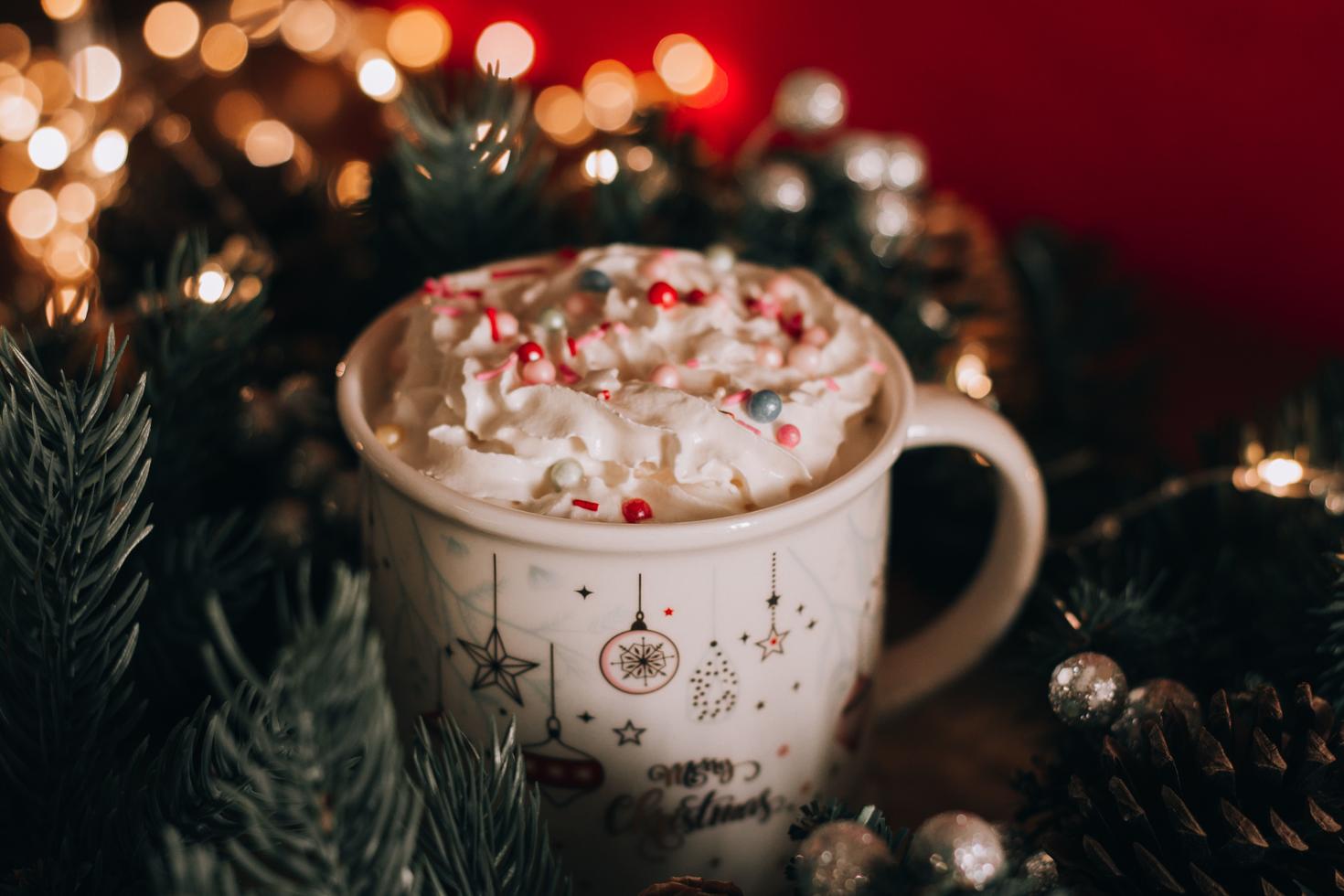 Mug garni de crème fouettée et de pépites de Noël, entouré de feuilles de pin, de pommes de pin et de guirlandes lumineuses.
