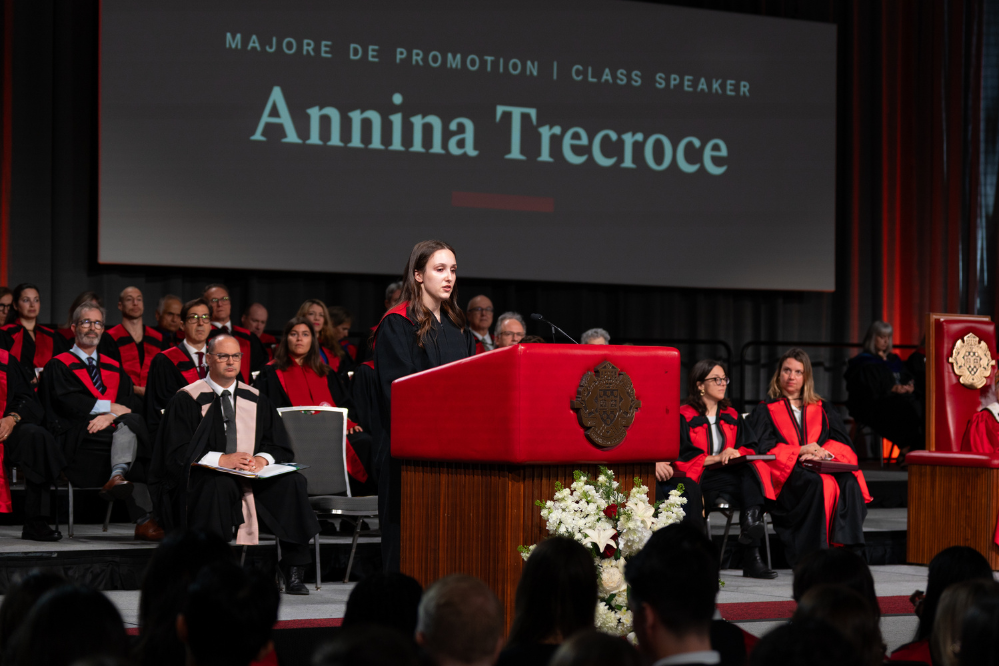 Une jeune femme, vêtue d’une toge noire, se tient à un podium. Derrière elle, de nombreuses personnes sont assises, elles aussi vêtues de toges noires avec des épitoges rouges.
