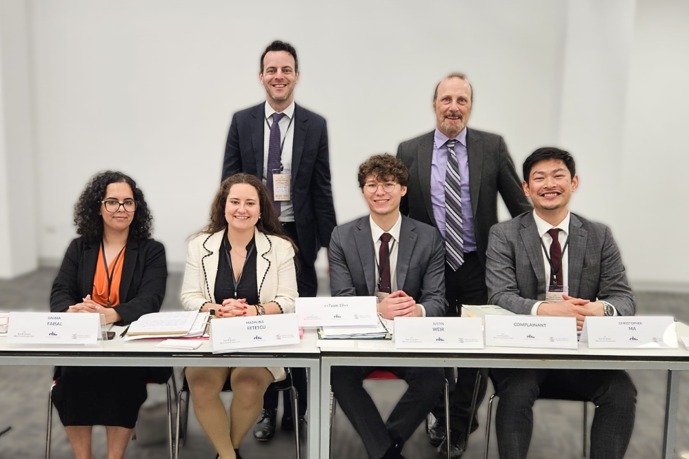 Five university students, 2 women and 2 men, sit at a large desk with books in front of them. Behind them, two men in suits stand.