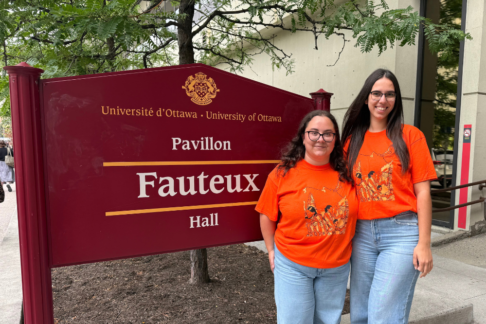 Deux femmes, vêtues de chandails orange, se tiennent près de l'enseigne du pavillon Fauteux.