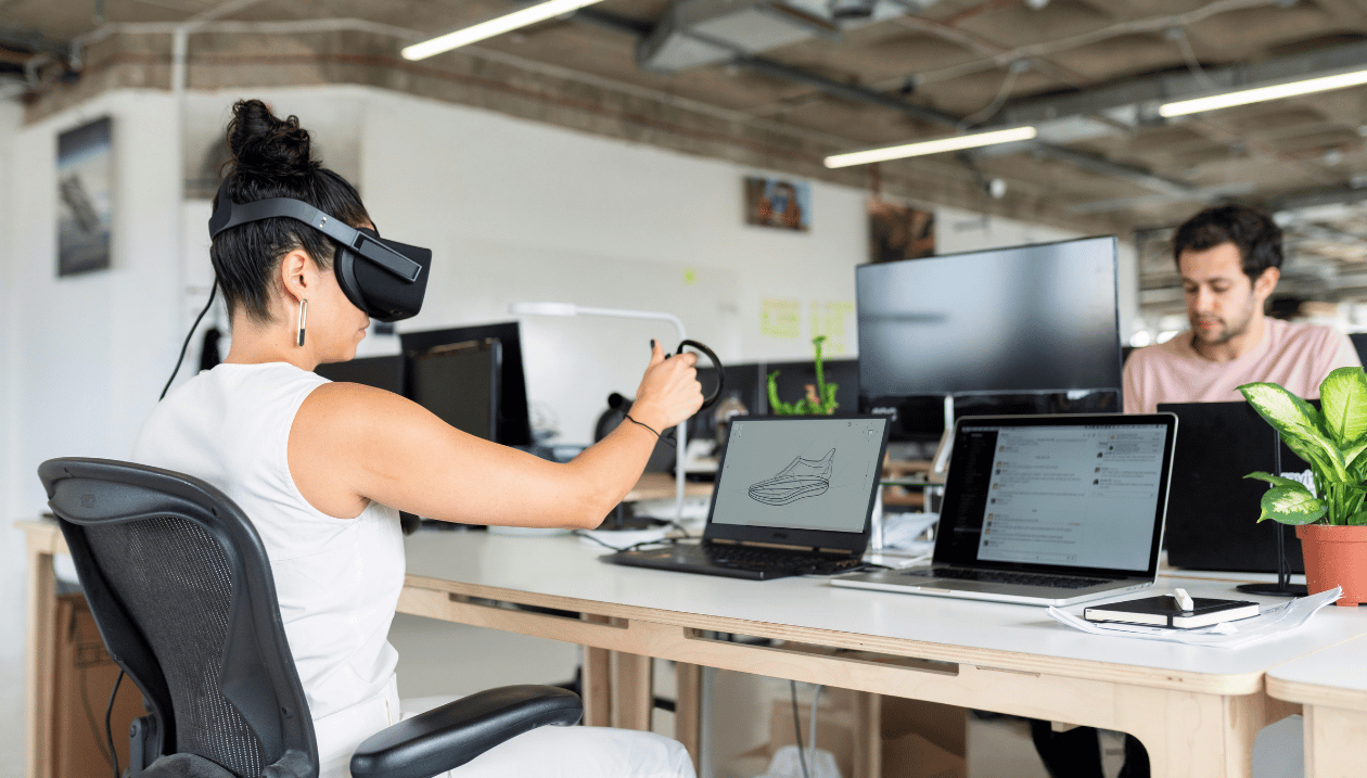 Person using a virtual reality headset at a desk, with a laptop displaying a 3D shoe model.