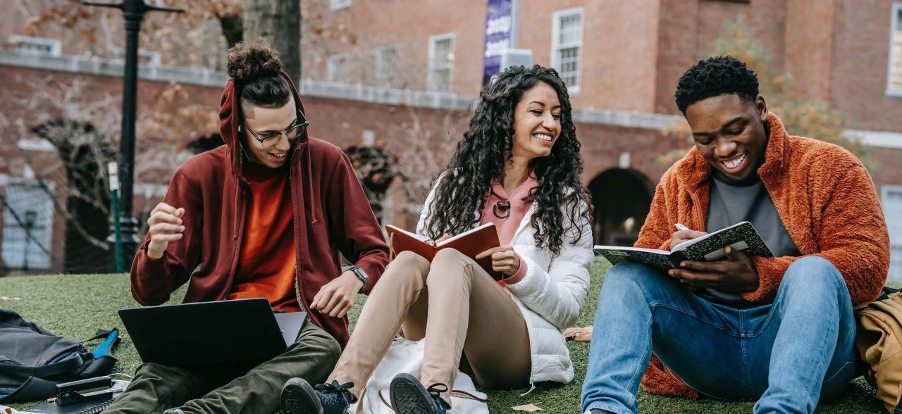 Groupe de trois étudiants noirs assis sur la pelouse devant le bâtiment du campus avec leurs cahiers et ordinateurs portables.
