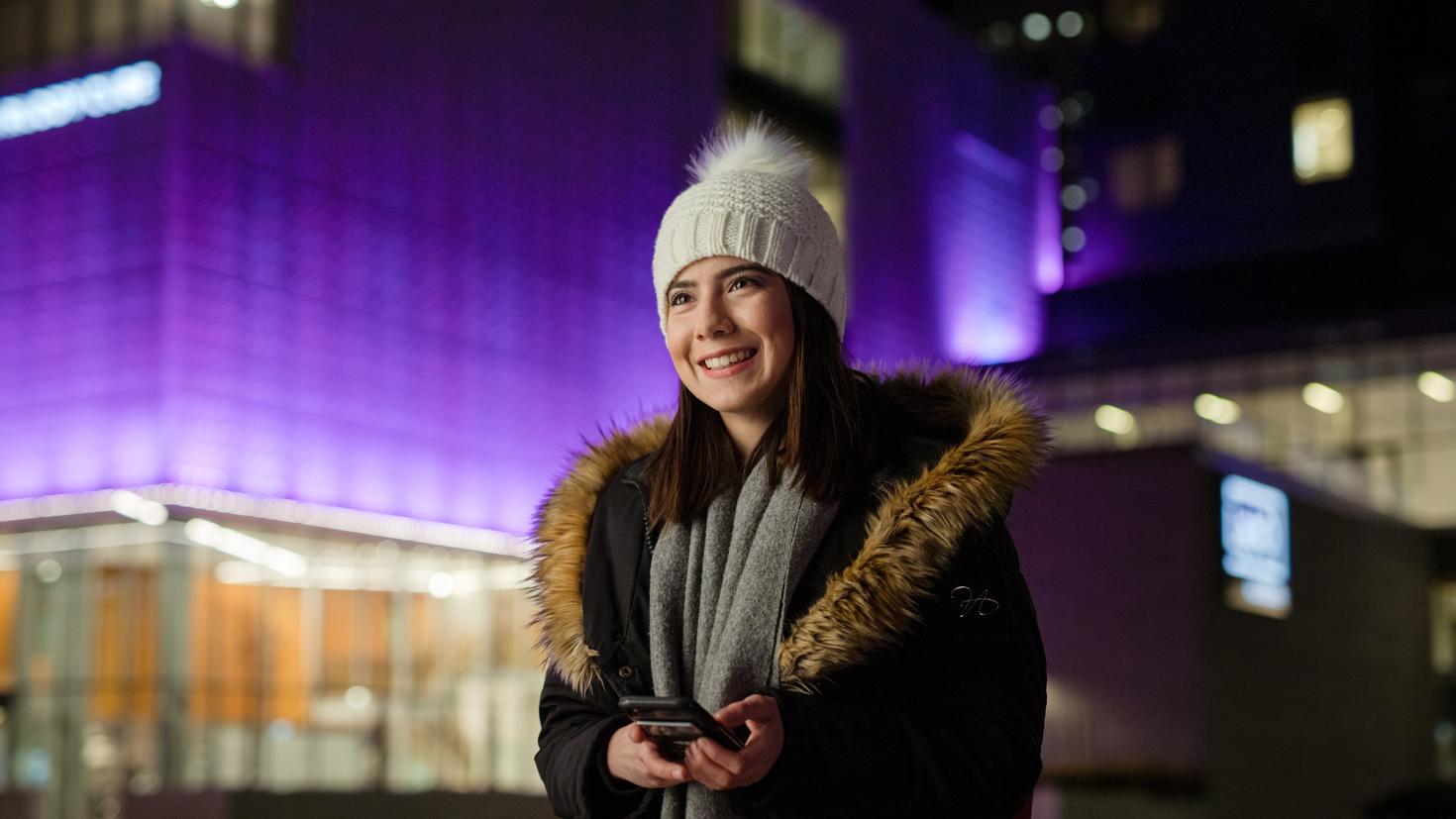 Une étudiante vêtue d’une tuque et d’un parka sourit sous un ciel nocturne, et figure en arrière-plan flou le centre-ville d’Ottawa tout illuminé, en périphérie du campus.