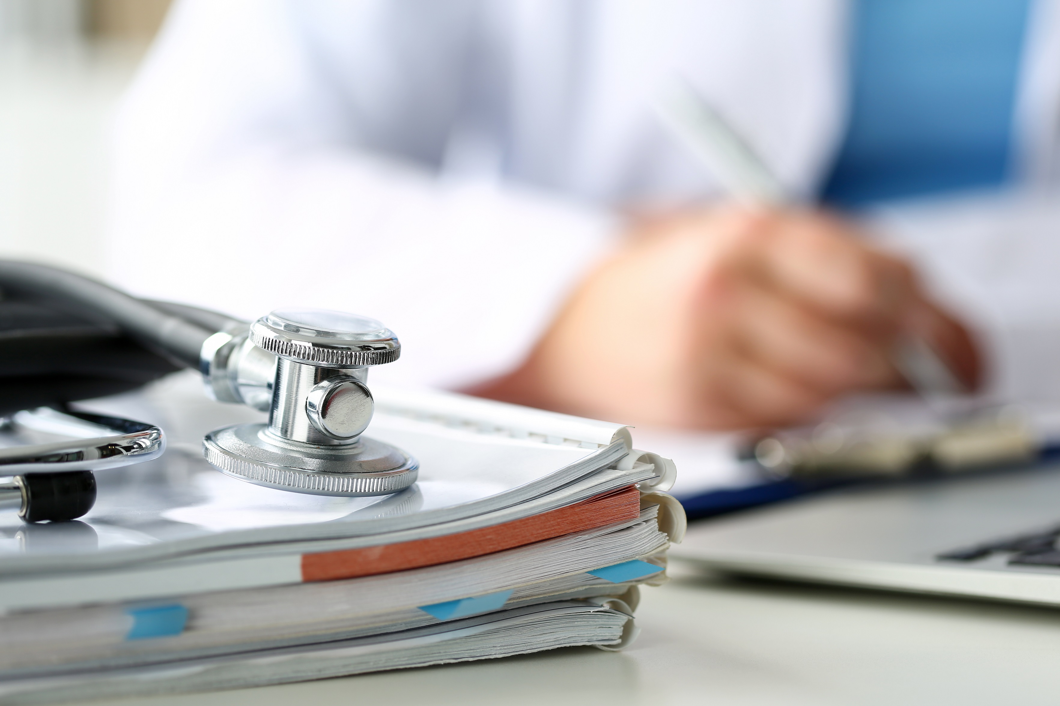 Tête de stéthoscope posée sur des formulaires médicaux sur une planchette à pince, en gros plan, tandis qu'un médecin travaille à l'arrière-plan.