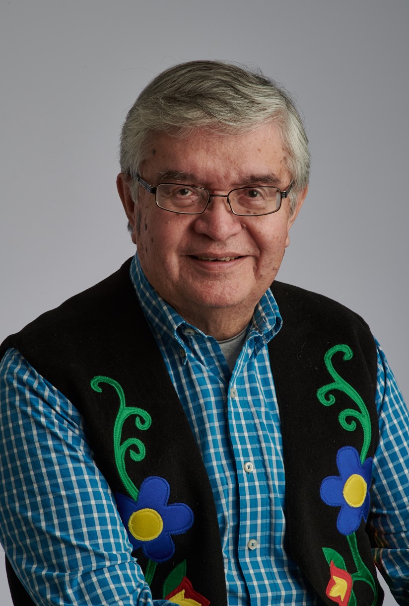 Un homme aux cheveux gris, portant des lunettes noires et une veste à motifs floraux d'inspiration autochtone, posant devant un fond beige-brun
