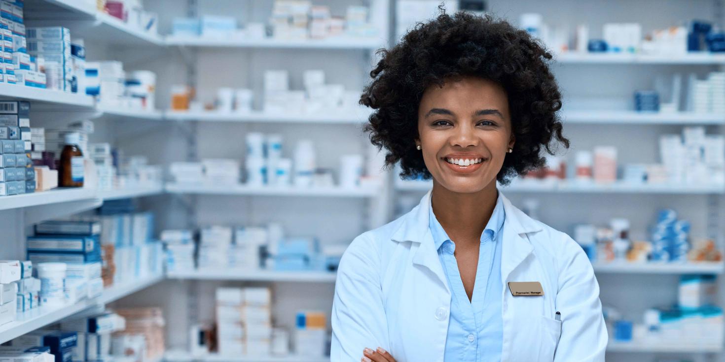 Pharmacienne souriante debout dans une pharmacie avec des étagères de médicaments derrière elle.