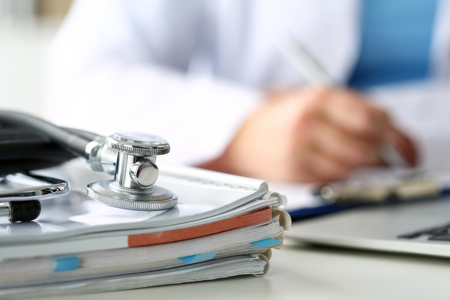 Stéthoscope posé sur des dossiers médicaux pendant qu’un professionnel de la santé écrit sur un porte-documents à l’arrière-plan.
