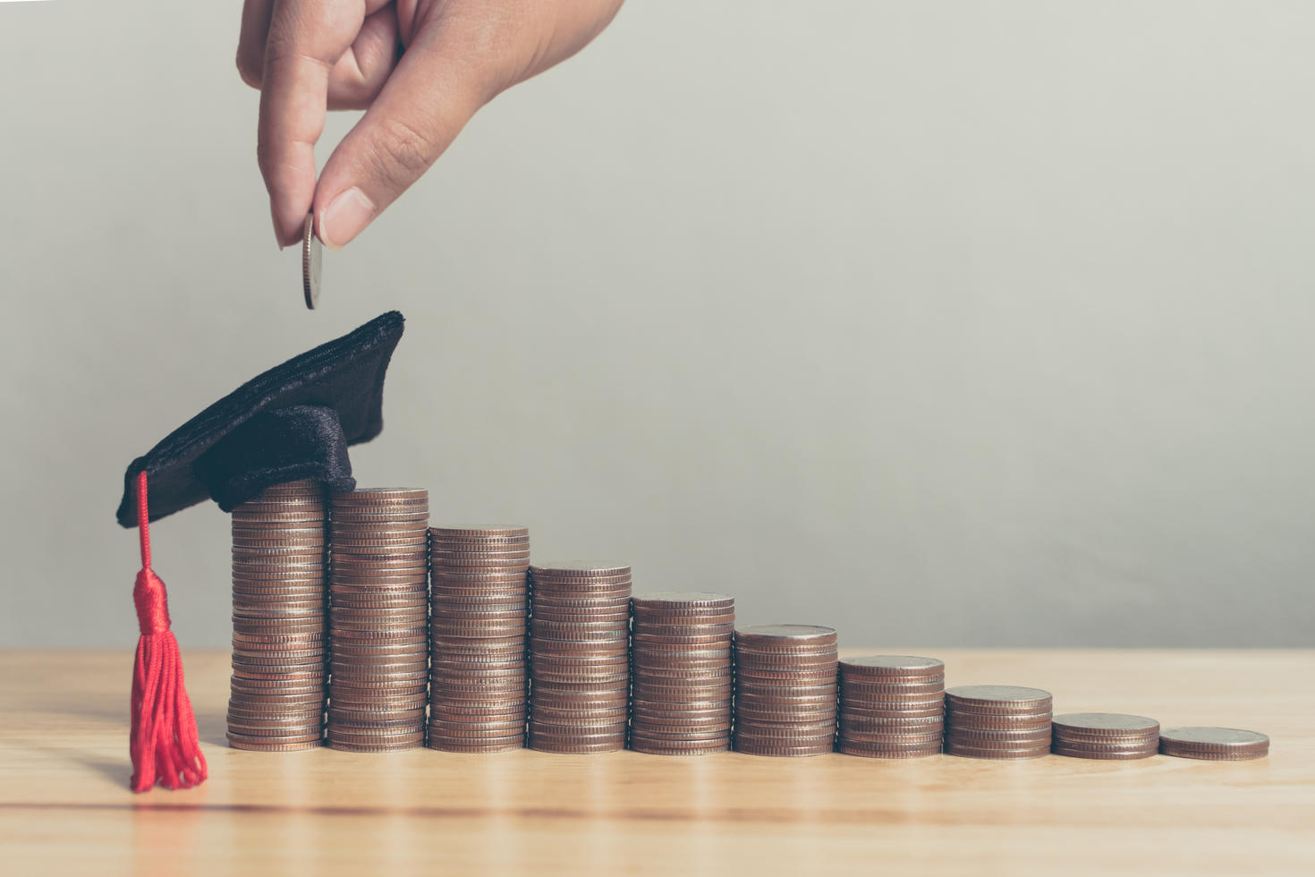 Une main dépose un penny sur une pile de pennies qui ne cesse de grandir, au sommet de laquelle trône un chapeau de diplômé.