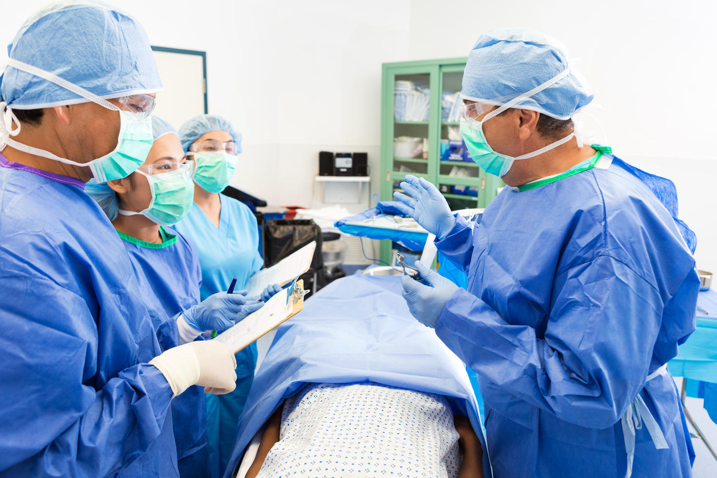 Un chirurgien adulte caucasien donne des cours à un groupe d'internes pendant l'opération d'une patiente à l'hôpital. Ils examinent le dossier de la patiente et la préparent à l'opération. Les chirurgiens portent des blouses, des bonnets, des masques et des gants stériles.