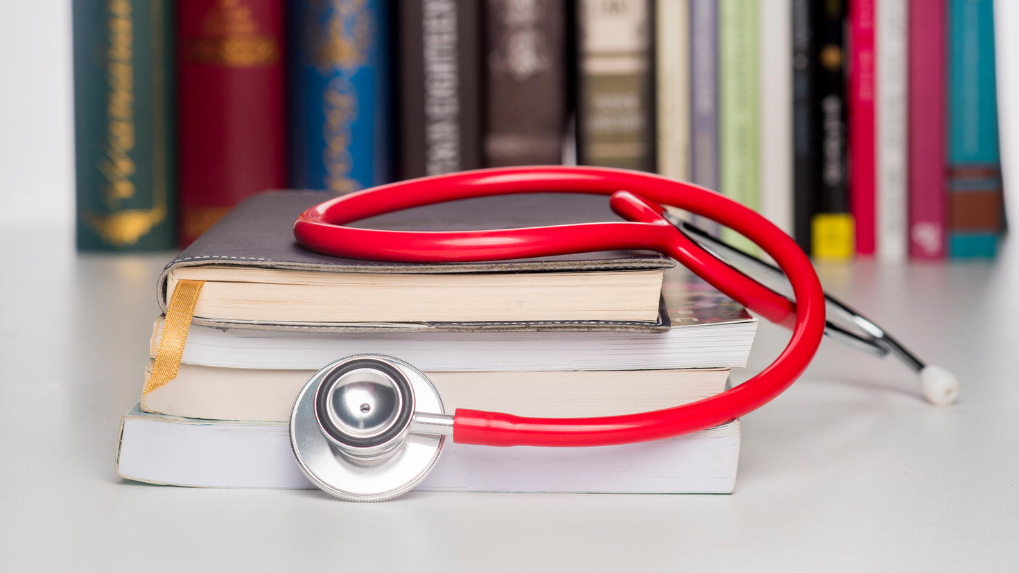 Gros plan d’un stéthoscope rouge posé sur une pile de livres devant une rangée de livres dans une bibliothèque.