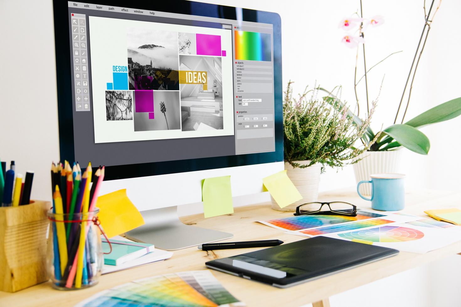 Un espace de travail lumineux équipé d'un écran d'ordinateur affichant un projet de design coloré avec diverses images et du texte. Des nuanciers de couleurs et des plantes en pot ornent le bureau.