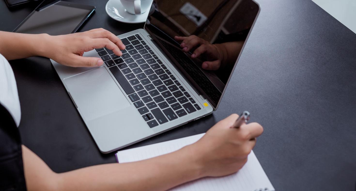 Des bras devant un ordinateur portable, prenant des notes sur un bloc-notes.