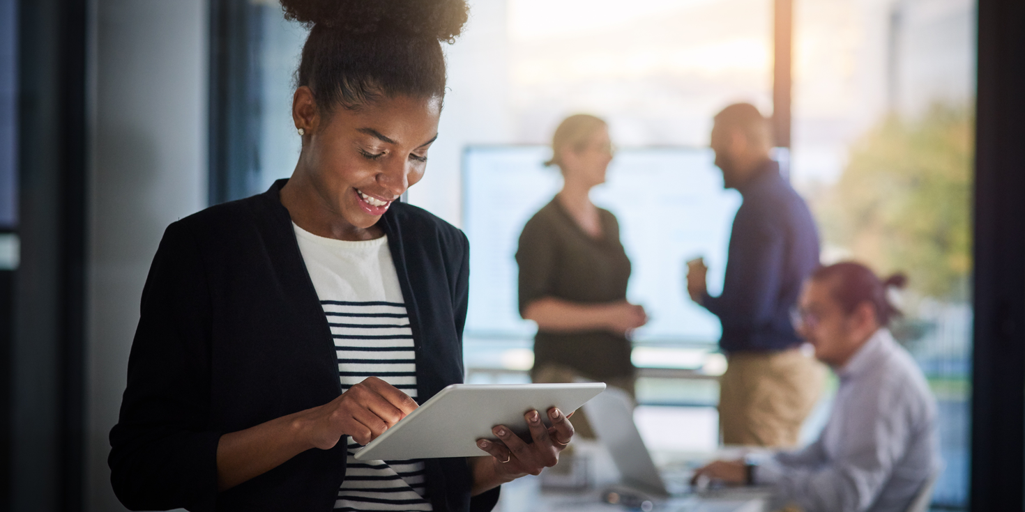 Une femme souriante utilise une tablette dans un environnement professionnel, tandis que des collègues discutent en arrière-plan.