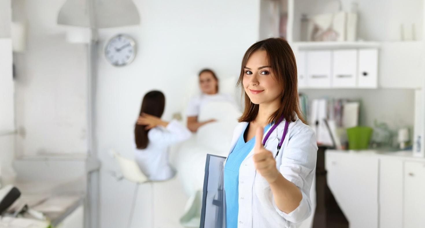 Une médecin souriante, tenant un bloc-notes et faisant le signe « OK » avec le pouce levé, lors de sa tournée dans le service.