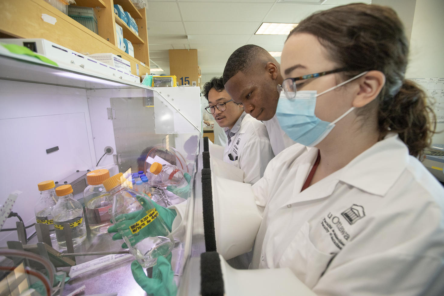 Trois étudiants portant des sarraus blancs manipulent des flacons sous une hotte de laboratoire dans un environnement de formation en sciences de la santé.